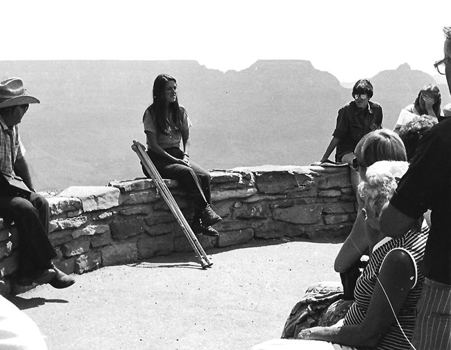 Eileen Szychowski in her NPS uniform sits on a low wall with her crutches at her side, addressing visitors. The Grand Canyon is in the background.