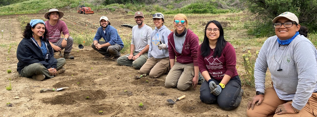 Members of Santa Monica Mountains National Recreation Area’s Restoration Program A diverse group of people sit and kneel on the ground in middle of field with small plants.