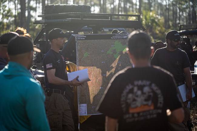 Several people stand around a map while a wildland firefighter gives a briefing.