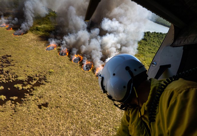 A man in protective gear looks out a helicopter window at the flames of a prescribed burn below