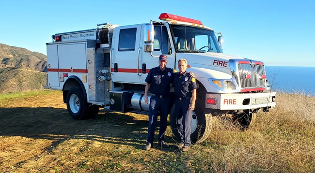 lt= Two men stand in front of a fire engine, with the ocean in the background