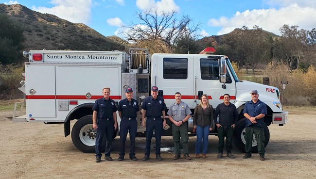 lt= A group of people stands in front of a fire engine in an outdoor setting
