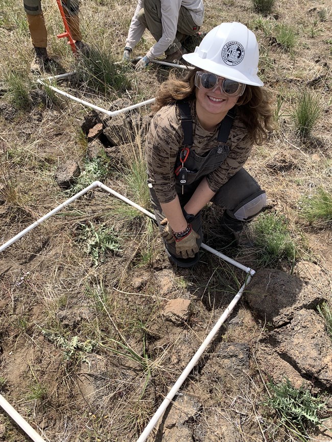 A young woman wearing a hardhat sits on the ground next to a rectangle made of PVC pipe.