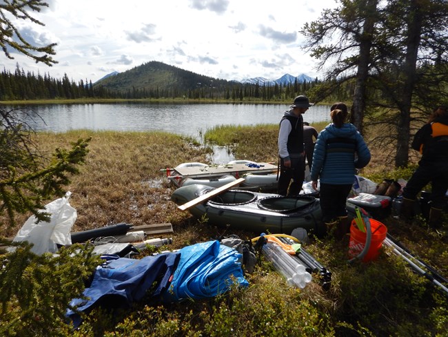 Three people stand near rafts and other gear by a lake surrounded by forest