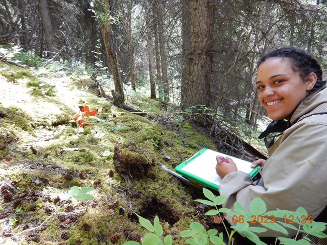 A woman holding a clipboard and kneeling in a forested area smiles at the camera