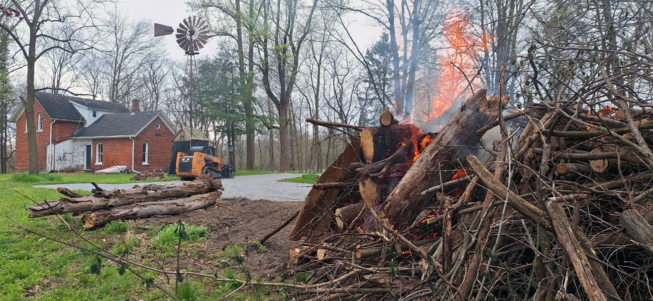 The Nelson House Pile Burn met goals of historic structure defensible space and cultural landscape management at Indiana Dunes National Park. Flames consume dead logs in a pile near a brick structure.
