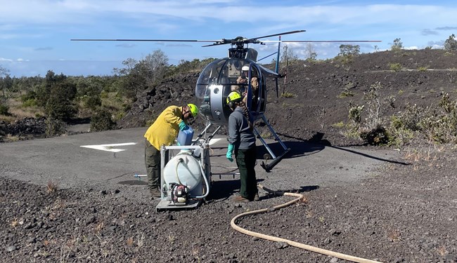 lt= Two people in front of a helicopter; one is pouring something from a container into a larger apparatus