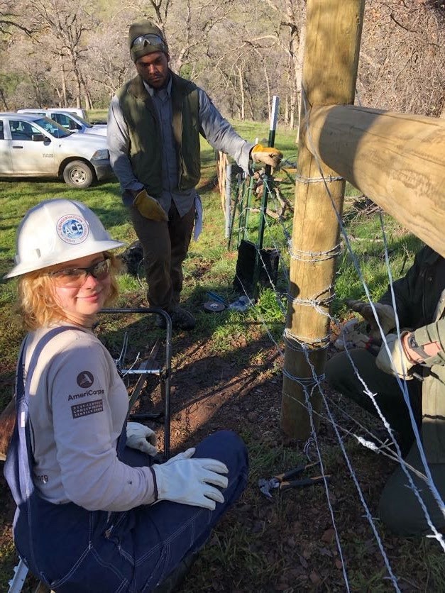 lt= A woman wearing a hard hat and gloves kneels in front of a fence smiling at the camera; a man stands in the background