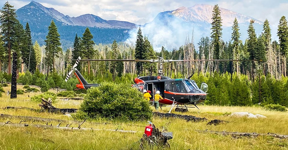 Helicopter 551 on the Red Fire. A crew works near a helicopter while smoke rises from a forest near the base of mountains in the background.
