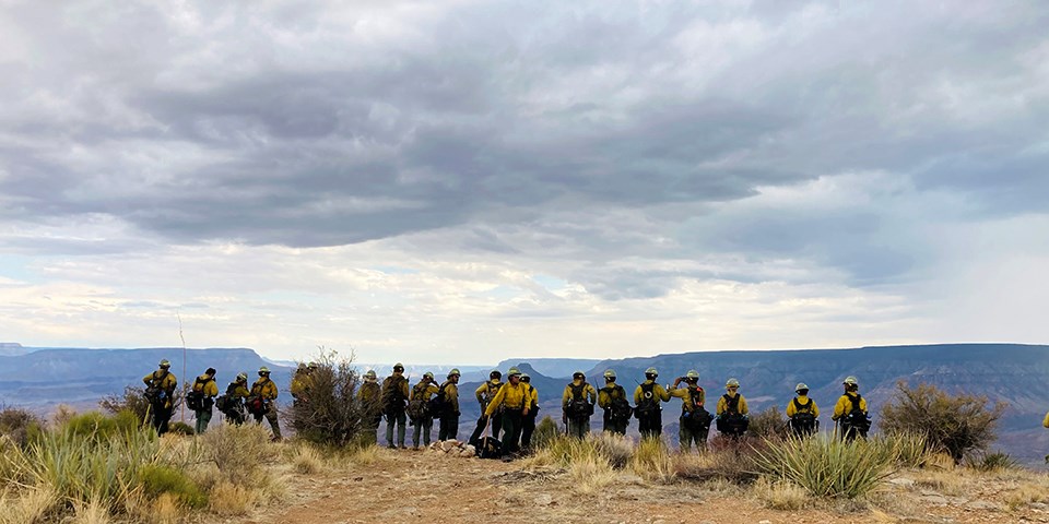 A line of firefighters stand on a ridge looking out into an extensive canyonland. A line of firefighters stand on a ridge looking out into an extensive canyonland.