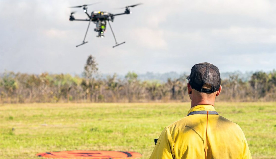 A person wearing Nomex and a baseball cap looks up towards a small uncrewed aircraft (UAS).