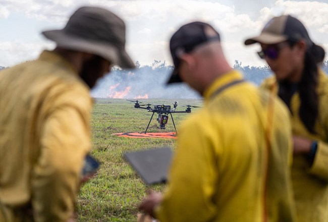 Three people look at a screen while an uncrewed aircraft is on a landing pad nearby and beyond that flames consume vegetation.