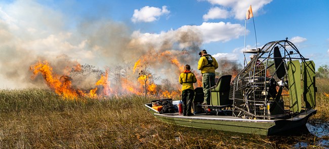 Two people stand in an air boat in a swampy area watching flames burning vegetation just ahead of them