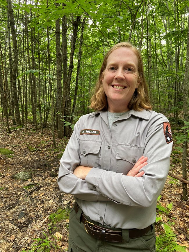 Kate Miller a woman in uniform crosses her arms and smiles at the camera with trees surrounding her