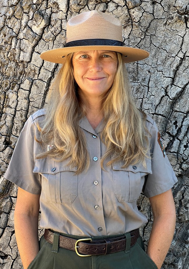 A tan-uniformed person with a wide-brimmed straw hat, long brown to blonde hair past their shoulders, smiling with blue eyes.