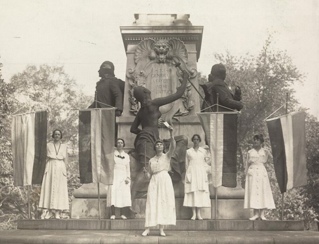 women stand with flags in front of a statue of Lafayette LOC