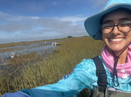 Natalie wears a sun hat and sun protective clothing while taking a selfie in front of a marsh.