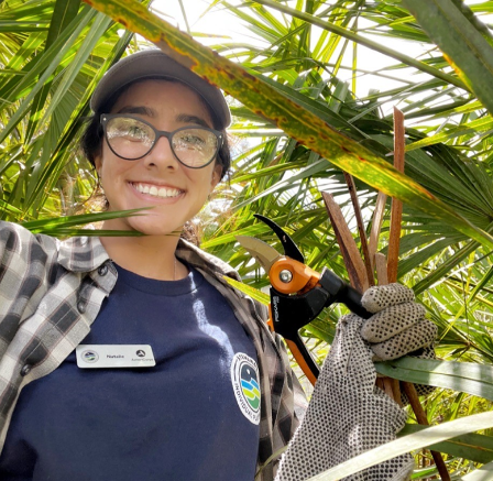 Natalie holds a set of pruning shears while surrounded by palm fronds.