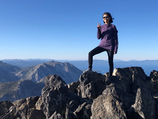 Woman stands on top of rocky peak, making a victory sign with her right hand.