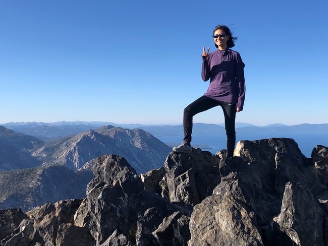 Natalie Kim Woman stands on top of rocky peak, making a victory sign with her right hand.