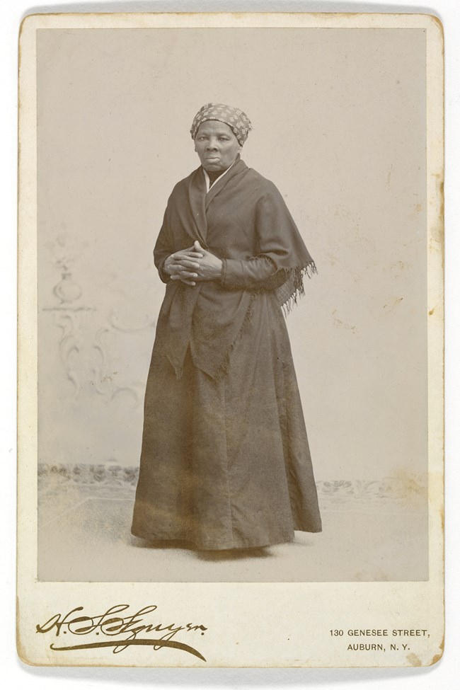 Harriet Tubman stands stoically in front of a plain background. She wears a plain dress and shawl, and a patterned headwrap.