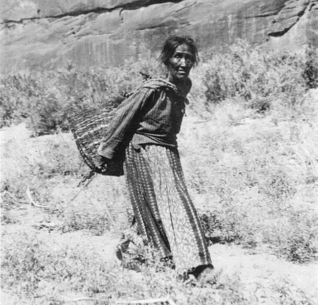 Black and white picture of a Navajo woman in a dress , carrying a wickerwork basket with peaches.