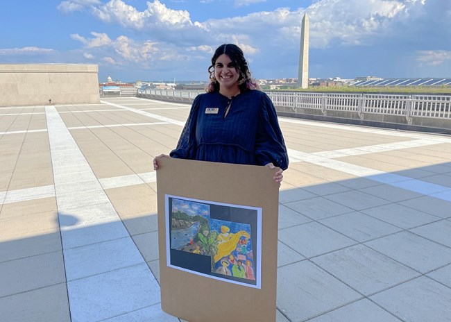 Nayelie Morales person holding a photo of two canvases painted with different trees and nature, as well as different people that depict Puerto Rican social justice leaders. The Washington Monument can be seen in the background