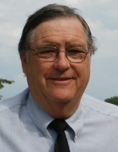 Photo of a man with glasses, wearing a light blue shirt and dark tie, standing outdoors with trees behind