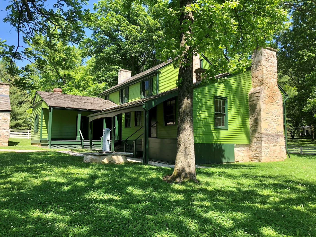 Green two story frame house surrounded by green trees and white fence.