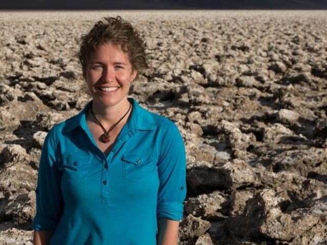 Woman facing camera smiling, standing in arid desert landscape.
