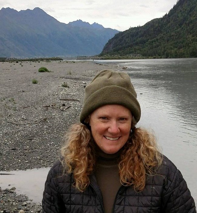 A woman in a hat and jacket with a river and mountains behind her.