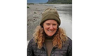 Nina Chambers Portrait of Chambers in an olive green beanie and a black puffy jacket by a river, smiling for the camera.