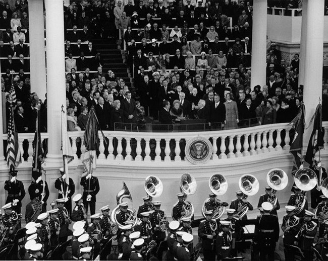 Military band plays at Nixon's inauguration in January 1973 A uniformed military band stands in rows, with a line of brass players standing against a white building, flanked by men carrying flags.