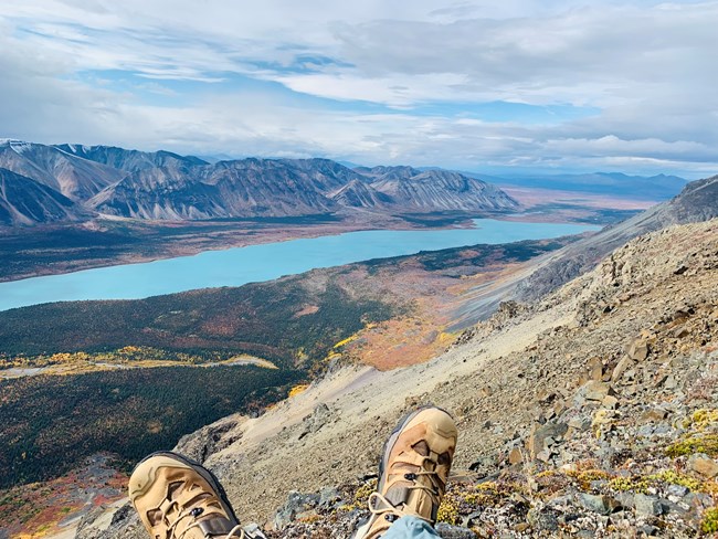 Noah Van Lith Hike Hiking boots in the foreground. Vantage point view overlooking a blue lake surrounded by trees an mountains. Trees are green yellow, and red.