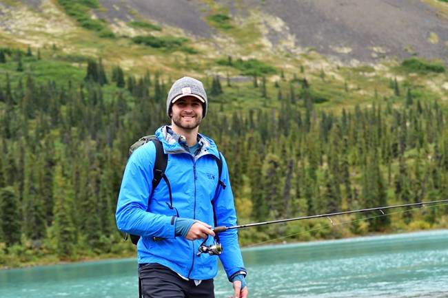 Noah Van Lith2 A man wearing a blue jack carrying a backpack stands in front of a turquoise-blue lake surrounded by trees.
