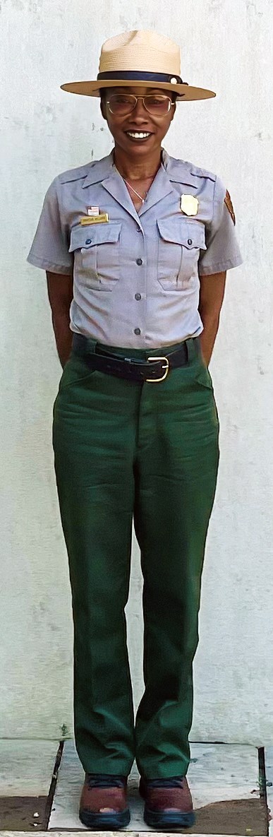 Smiling woman in NPS uniform, gray shirt, green pants, badge, straw hat with hands folded behind her back