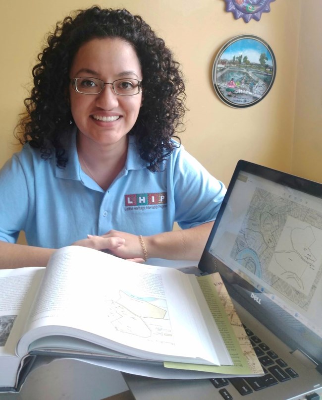 Young woman in uniform polo sits at table with open book of maps and computer