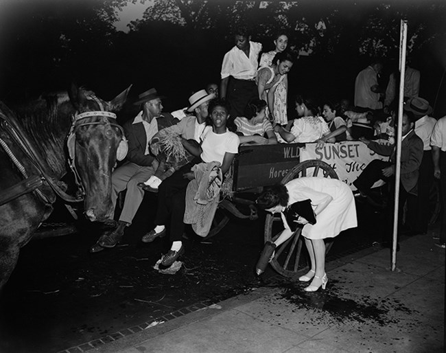 Black and white photo. A large group of African American men and women sitting and climbing into a wooden horse-drawn wagon. They are dressed up for an evening on the town. Left: a dark colored horse, perhaps waiting for another wagon-load of revelers.