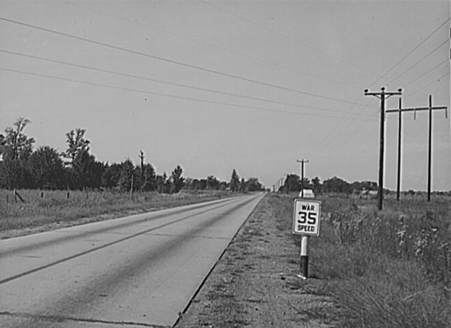 Black and white photo looking down a paved road with fields and trees. On the side of the road is a speed limit sign.