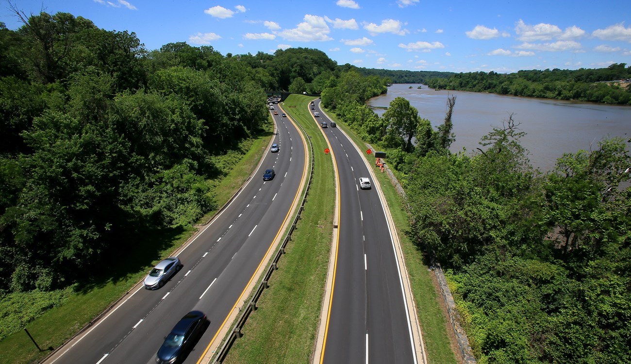 Looking down on cars traveling on two paved highways, through a green forested area.