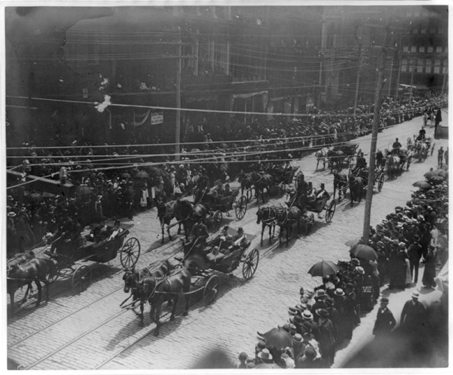 Black and white birds eye view of a funeral procession taking place with people inside multiple carriages pulled by horses