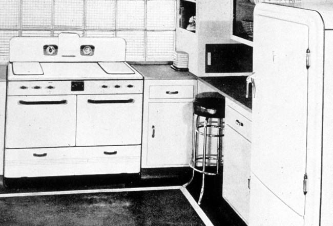 Black and white photo of a kitchen with enamel stove, white cupboards, a refrigerator, and a stool.
