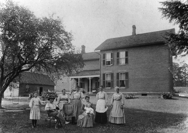 A man, a girl, five women, and a baby pose in front of a large brick house and an outbuilding.