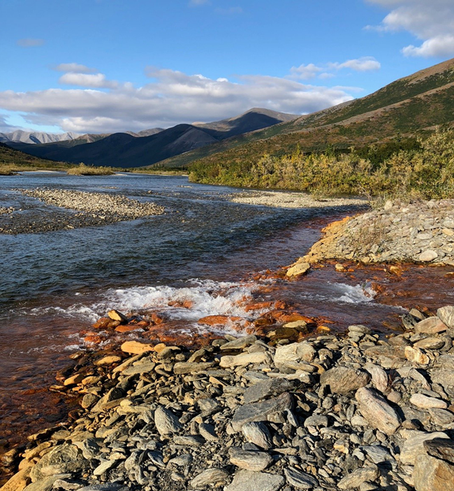 image of a headwater monitoring site in the Akillik River turned orange in response to iron release from permafrost soils. Mountains in the background under a partly cloudy sky.