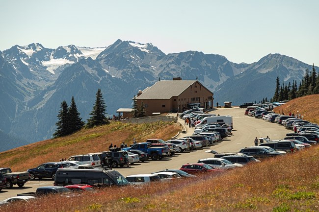 A house/lodge sits behind a parking lot full of cars on a sunny day. There is a mountain range in the background of the photo