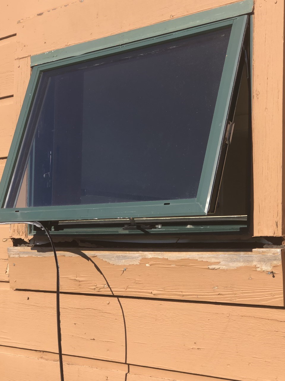 A dark green window with peeling tan paint, rotting trim and black telecommunication line.