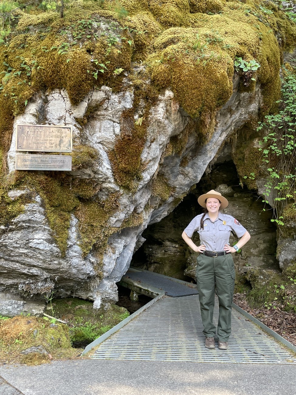 Ranger Sarah Sherwood at Oregon Caves A woman with a brown braid wearing the NPS uniform stands smiling with her hands on her hips. She is in front of a mossy rock wall on a metal walkway that leads away behind her into a crack in the mountain that is Oregon Caves.