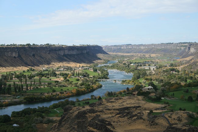 A snaking river makes its way through a valley bottom dotted with buildings and roads.