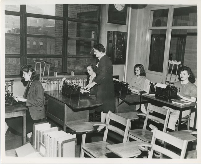 Black and white photo of a woman, center, helping a younger woman who is seated at a desk and using a typewriter. Three other young women are also seated at desks using typewriters. Two pairs of crutches lean against the wall beside two of the students.