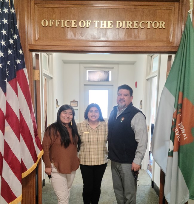 NPS Director A photo of three people a National Park Service intern, director assistance, and the director all standing by each other under the Office of the Director sign.